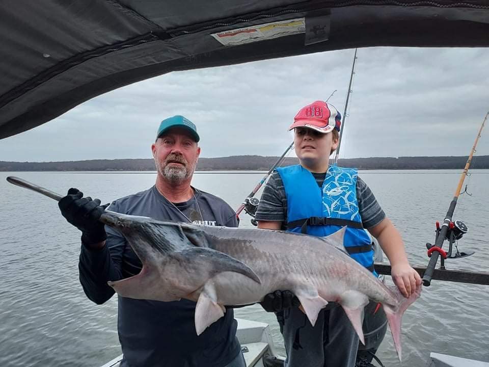 A guide and a young angler in a blue life jacket holding a paddlefish together under the boat canopy.