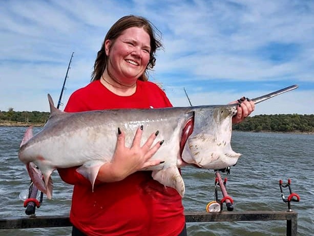 Smiling angler in a red shirt holding a large paddlefish across both arms, rods in holders and open water behind her.